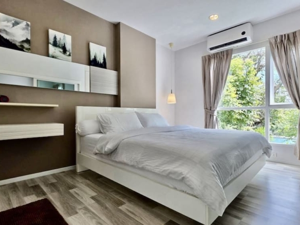 Modern white bed with white linens in a bedroom with a brown accent wall, floating shelves, and a large window with beige curtains.