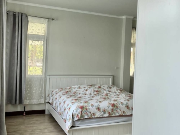 White bedroom with a floral duvet on a white bed against a pale wall, light wood floors, and gray curtains by the window.