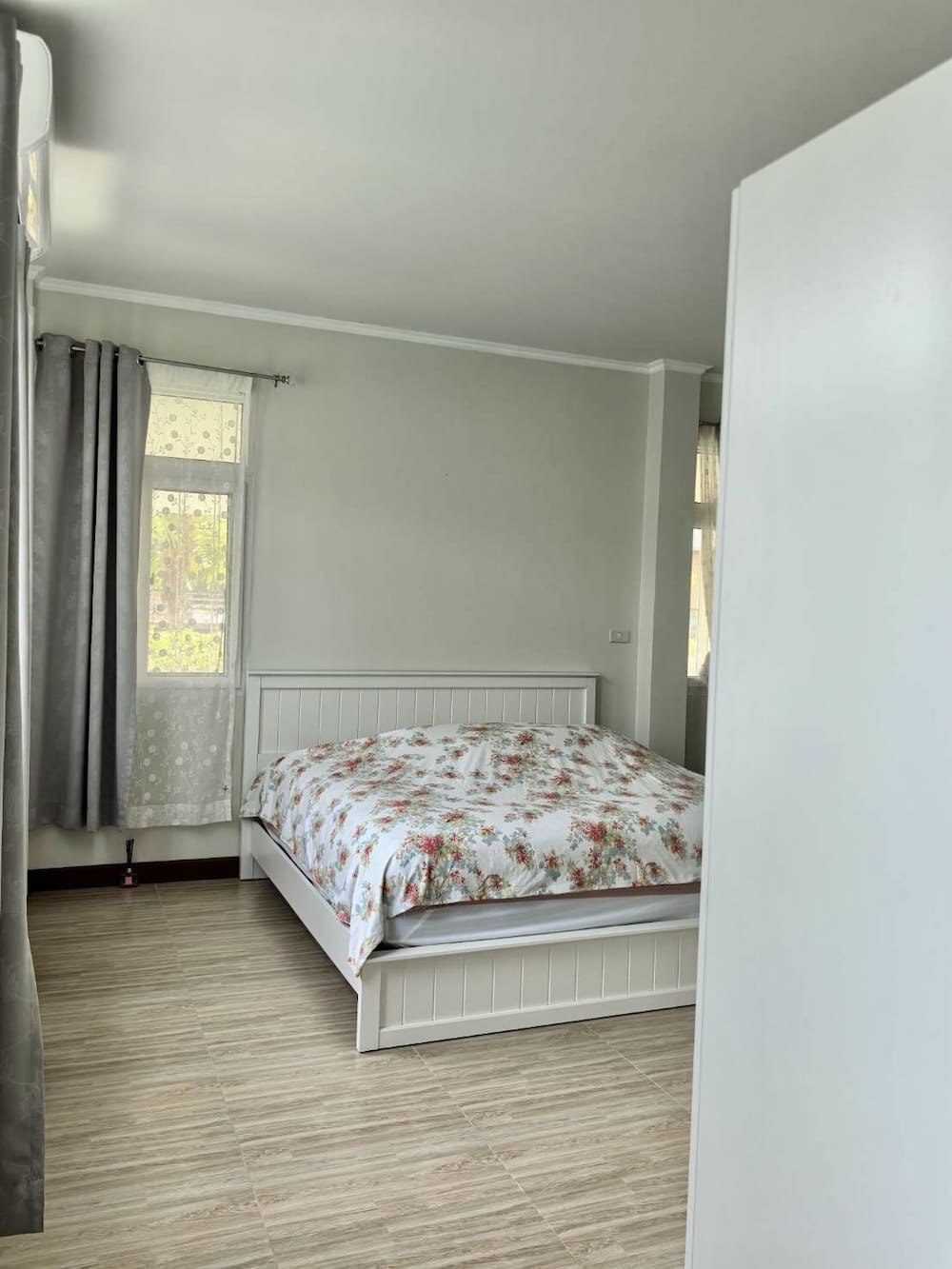 White bedroom with a floral duvet on a white bed against a pale wall, light wood floors, and gray curtains by the window.