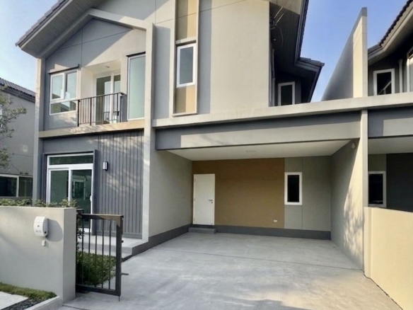 Modern two-story house with a covered carport, spacious concrete driveway, and a gated entrance; beige and gray exterior with a small balcony and sloped roof.