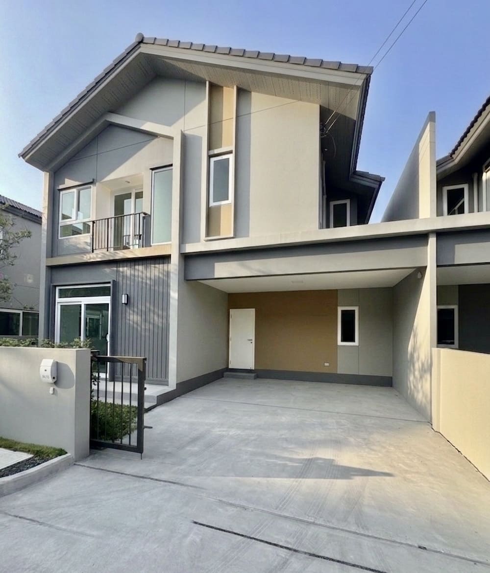 Modern two-story house with a covered carport, spacious concrete driveway, and a gated entrance; beige and gray exterior with a small balcony and sloped roof.