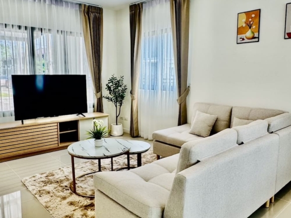 Bright living room with an L-shaped beige sofa, round marble coffee table, and a flat-screen TV on a wooden console.