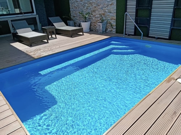 Outdoor pool with wooden deck, lounge chairs, and potted plants along a stone wall backdrop as sunlight shines on the water surface.