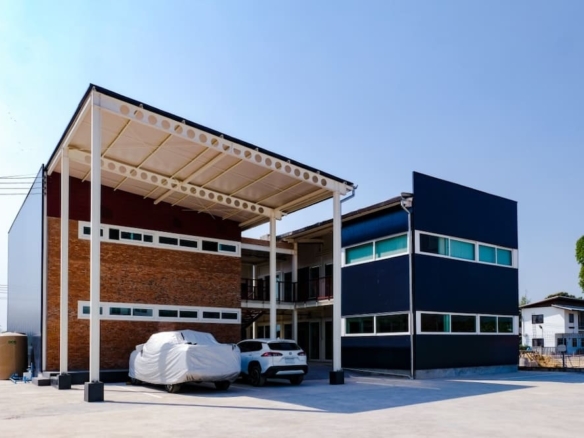 Modern two‑story office building with a white canopy, brick left wall and dark blue right section; two cars parked outside (one covered).