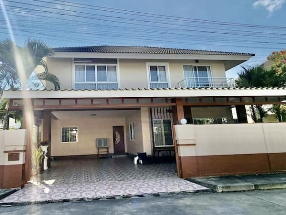 Two-story beige house with a covered carport, tiled driveway, and palm trees under a bright sky.