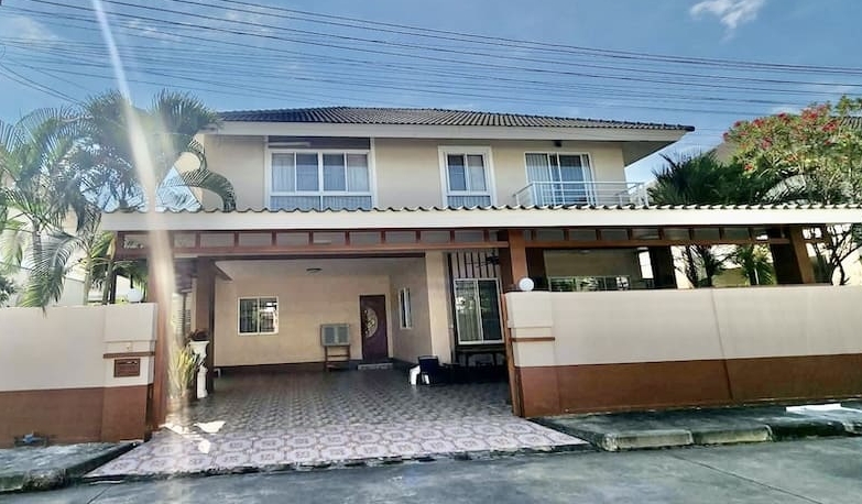 Two-story beige house with a covered carport, tiled driveway, and palm trees under a bright sky.