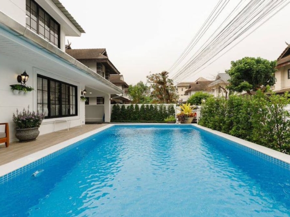 Rectangular blue swimming pool in a modern backyard beside a white house, with a wooden bench and potted plants along a wooden deck.