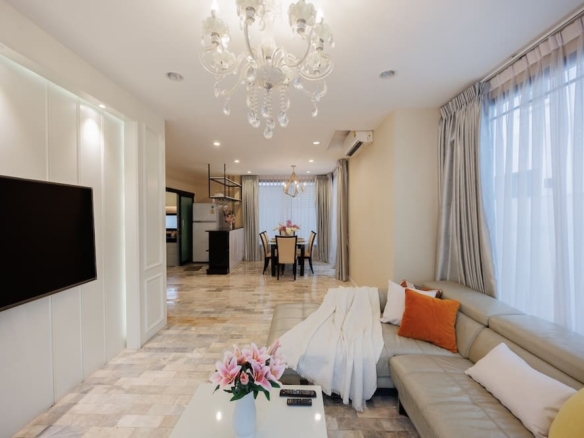 Luxurious open-plan living room with a chandelier, beige sectional sofa, orange and white cushions, and a TV on the wall.