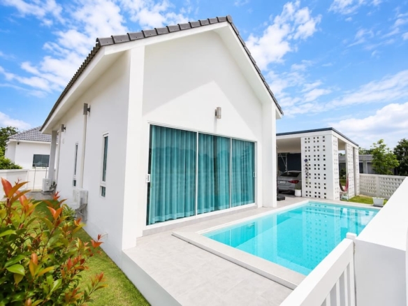 White modern house with a rectangular blue-tiled pool in the foreground and a carport to the right on a sunny day