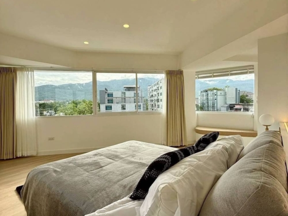 Bright bedroom with panoramic city and mountain views through large windows, white bedding, and a gray throw on the bed.