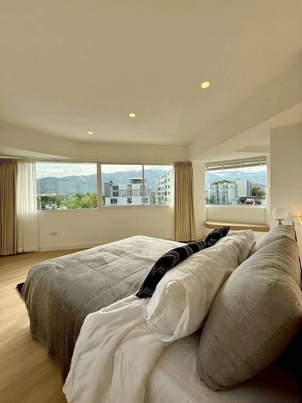 Bright bedroom with panoramic city and mountain views through large windows, white bedding, and a gray throw on the bed.