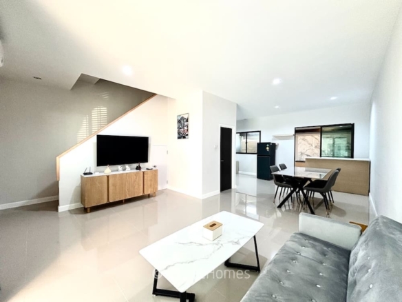 Bright open-plan living and dining area: gray tufted sofa, marble coffee table, TV on a wooden console, staircase nearby, and a dining table with chairs.