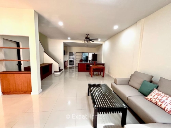 Spacious open-plan living room with beige sofa, black coffee table, and cushions, leading to a wooden dining area and kitchen in the background.