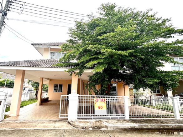 Two-story beige house with tiled roof, a covered carport, and a large green tree behind a white gate.