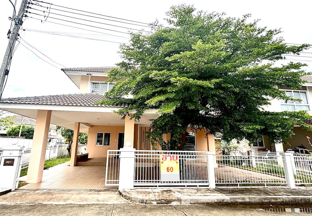 Two-story beige house with tiled roof, a covered carport, and a large green tree behind a white gate.