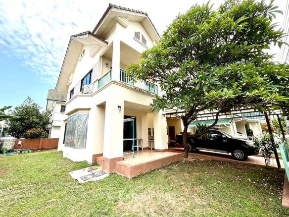 Two-story beige house with a covered porch, green balcony rails, and a large tree in the front yard with a black SUV under a carport.