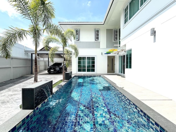 White two-story house with a rectangular tiled pool in a sunny backyard, palm trees, and a parked car under a carport to the left.