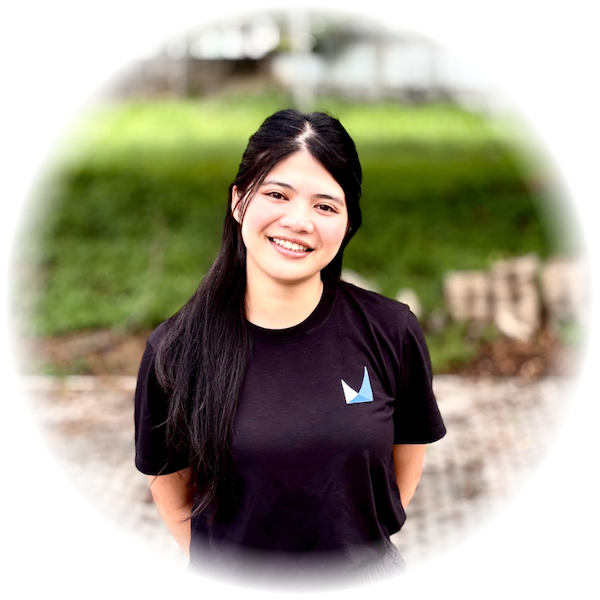 Smiling woman with long dark hair wearing a black t-shirt outdoors, circular vignette background.