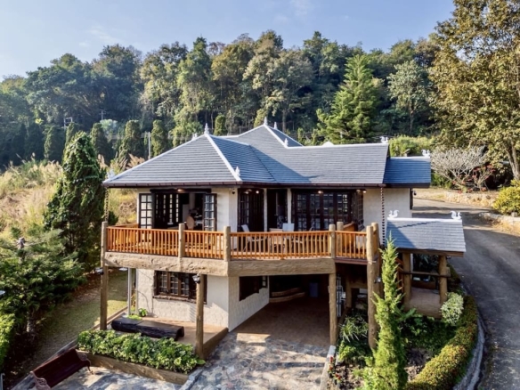 Two-story house with a wooden balcony and grey tiled roof, surrounded by trees and a curved driveway to the right