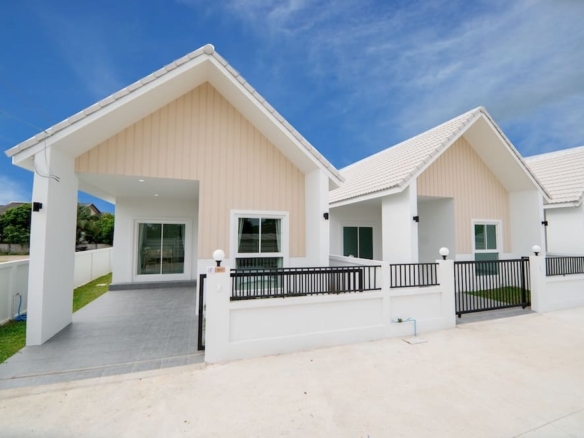 Row of modern single-story homes with peaked roofs, beige siding, white accents, and black railings along a gated walkway set under a bright blue sky.