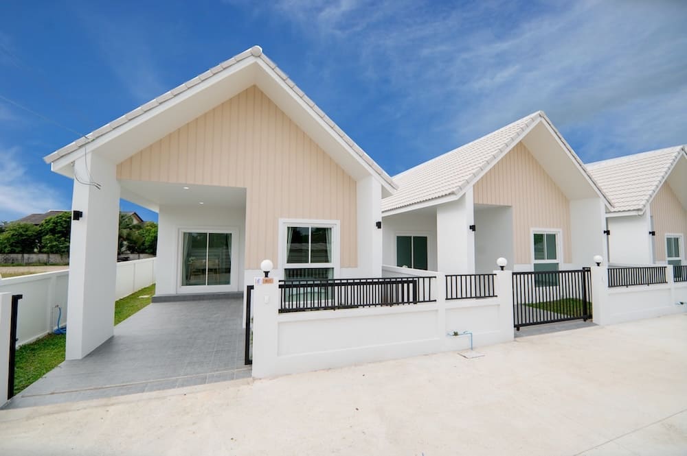 Row of modern single-story homes with peaked roofs, beige siding, white accents, and black railings along a gated walkway set under a bright blue sky.