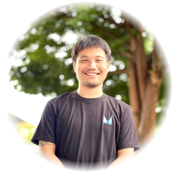 Smiling man in a black t-shirt with a small blue logo, outdoors with trees in the background, circular vignette.