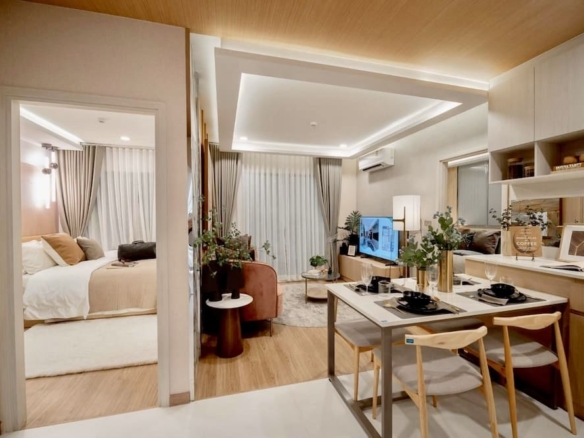 Open-plan living area with a dining table for two, sofa, TV, and potted plants; doorway reveals a bedroom with a bed and beige bedding.