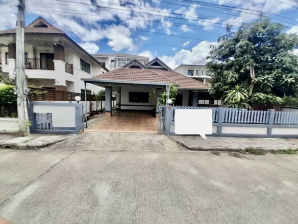 House with a brown-tiled roof and open carport, paved front yard, and blue fence along the street. Blue sky with clouds above.