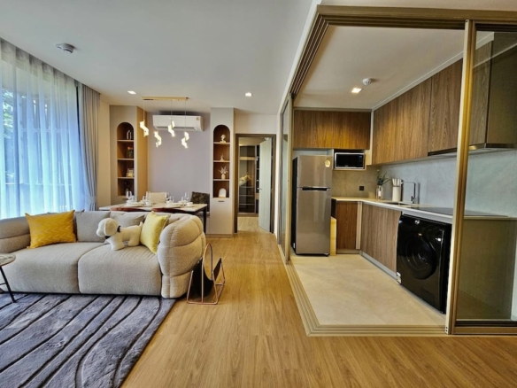 Open-plan living room and kitchen in a modern apartment: gray sectional with yellow cushions, a dining table, and built-in shelves with decorative items.