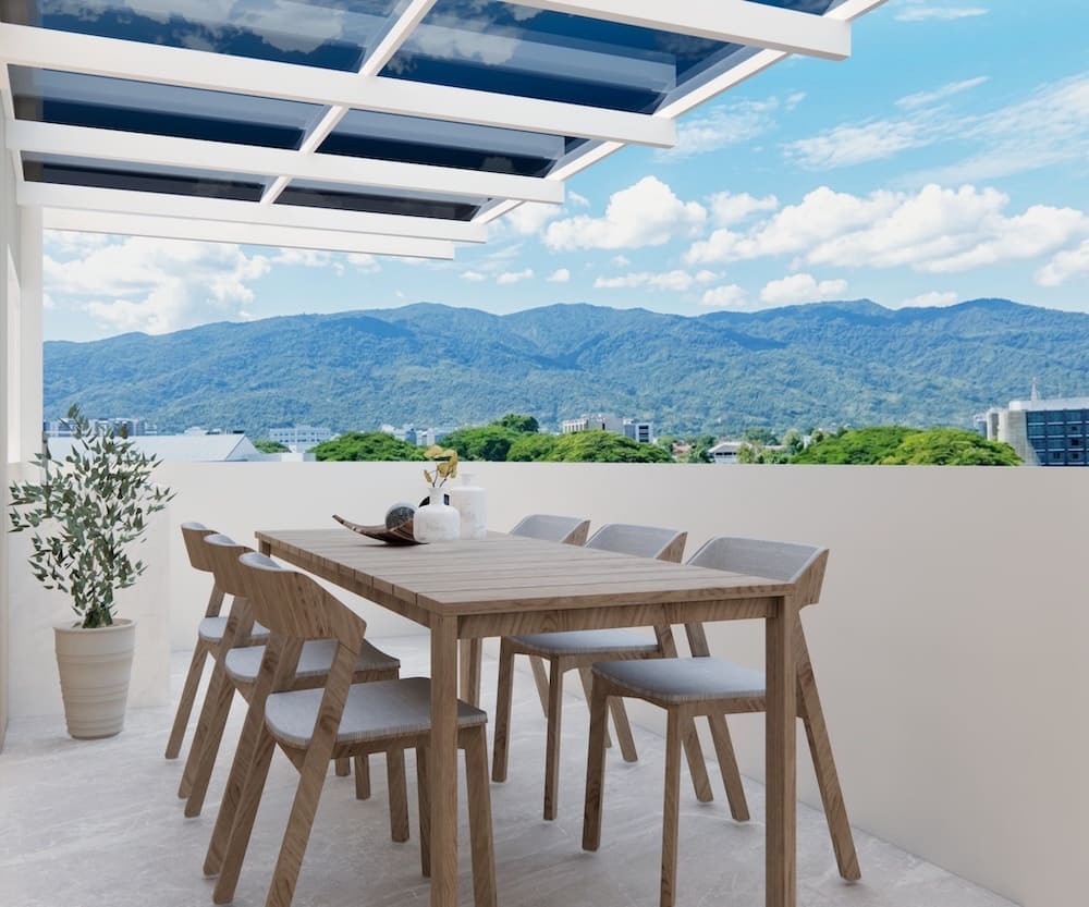 Rooftop patio with a wooden dining table and six chairs, potted plant, and a glass-covered trellis against a mountain landscape.