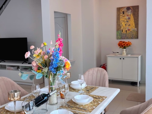 Modern dining area with a white table set for six, beige upholstered chairs, and a vivid flower centerpiece; TV and white cabinet in the background.