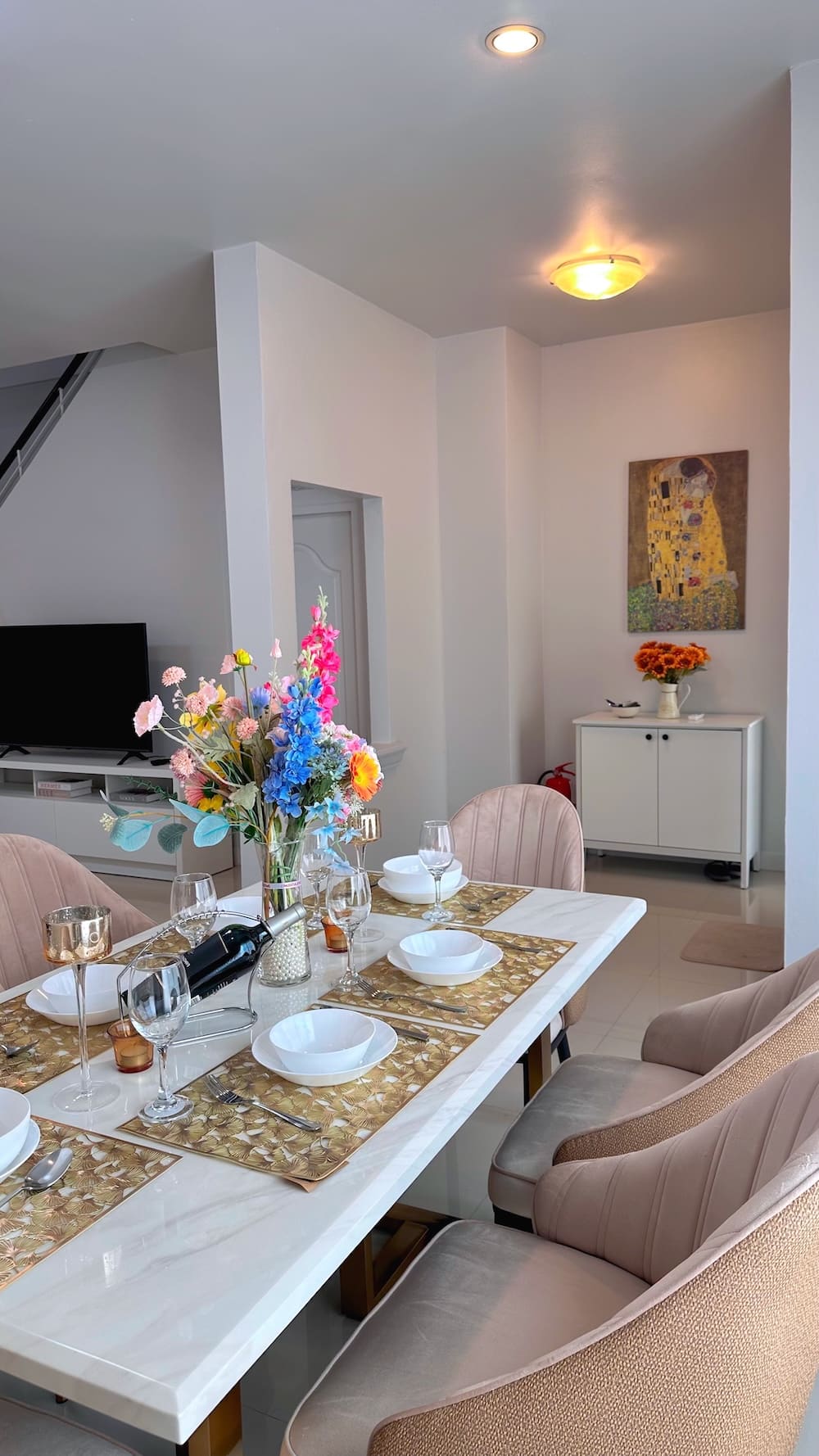 Modern dining area with a white table set for six, beige upholstered chairs, and a vivid flower centerpiece; TV and white cabinet in the background.