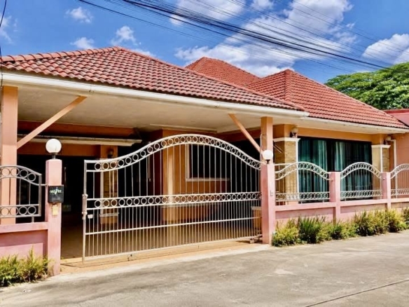 Front view of a single-story pink house with a red-tiled roof, enclosed by a decorative white metal gate and pink fence under a blue sky