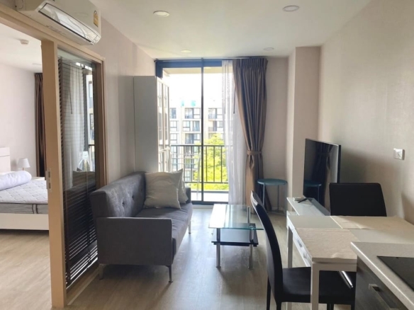 Modern studio living area with a gray sofa, glass coffee table, and a wall-mounted TV, opening to a balcony with sliding doors and city view at the end of the room.