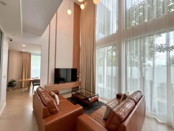 Bright living room with tall glass walls and sheer white curtains letting in natural light, brown leather sofa set, and a wall-mounted TV on a wooden panel.