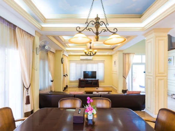 Bright dining table in the foreground with a bouquet, facing a living area with brown leather sofas and a wall-mounted TV under a decorative ceiling chandelier.