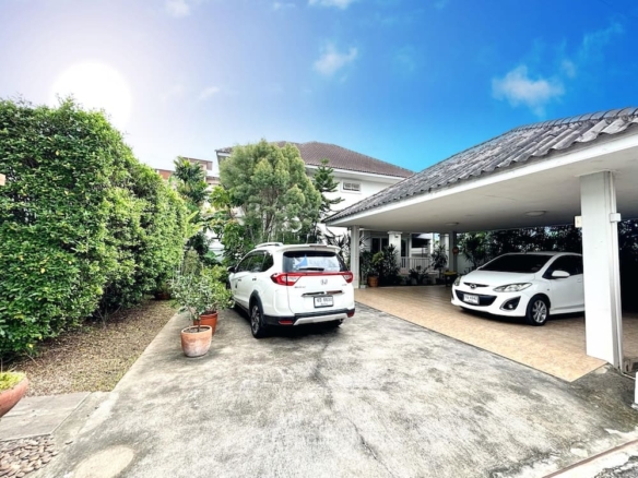 White SUV and white sedan parked in a residential driveway beside a covered carport, with green hedges and potted plants along the path.