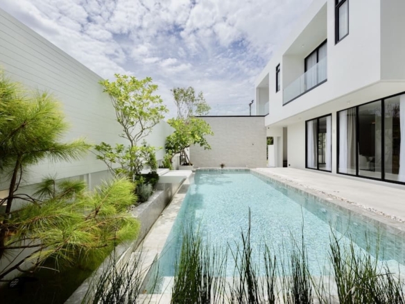 Modern white residence with a long rectangular pool, glass doors, and surrounding greenery under a blue sky.