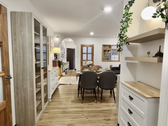 Bright open-plan dining area with light wood floors, white storage, and a green leafy arch above shelves near a dining table with grey chairs.