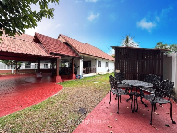 Wide view of a single-story house with red-tiled roof, open carport, and a black wrought-iron patio set on a red concrete patio.