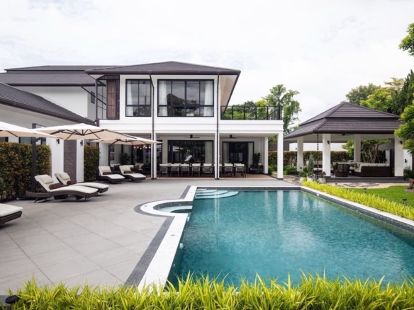 Contemporary two-story villa with a large rectangular pool, lounge chairs, and white umbrellas along a spacious patio.